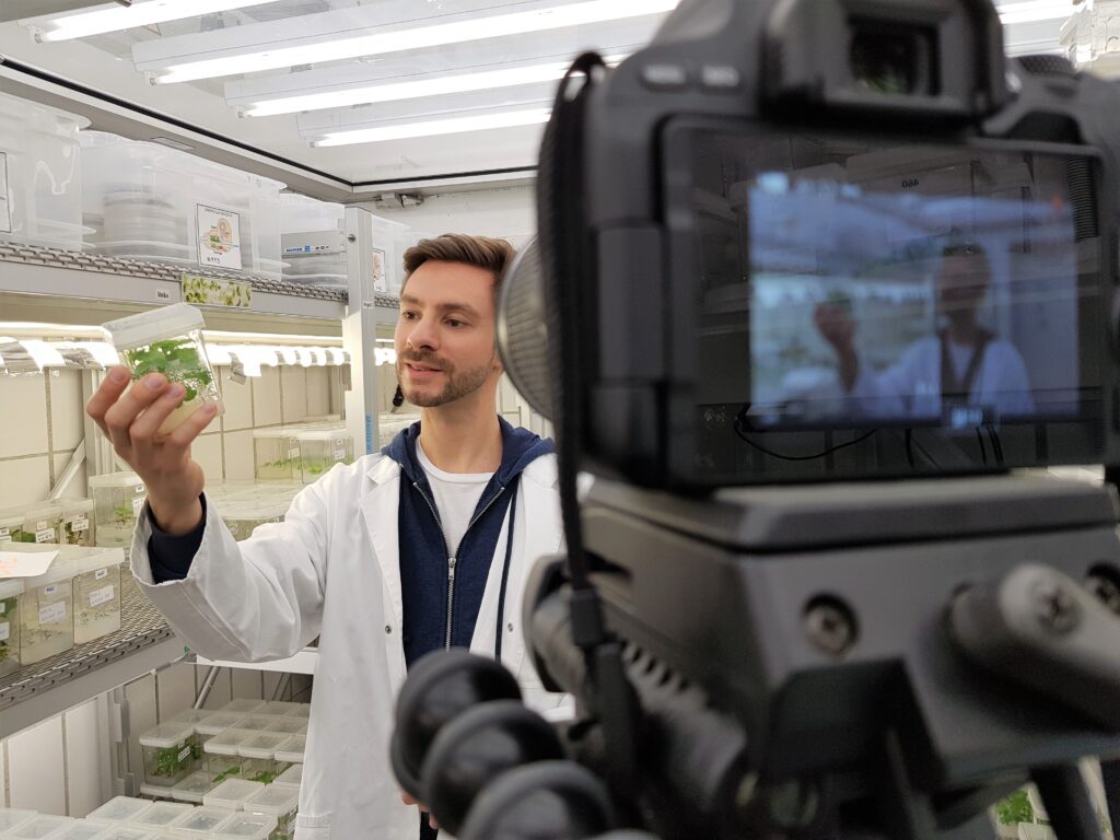 Wissenschaftler im Kittel vor der Kamera mit einer Pflanzenkultur in der Hand